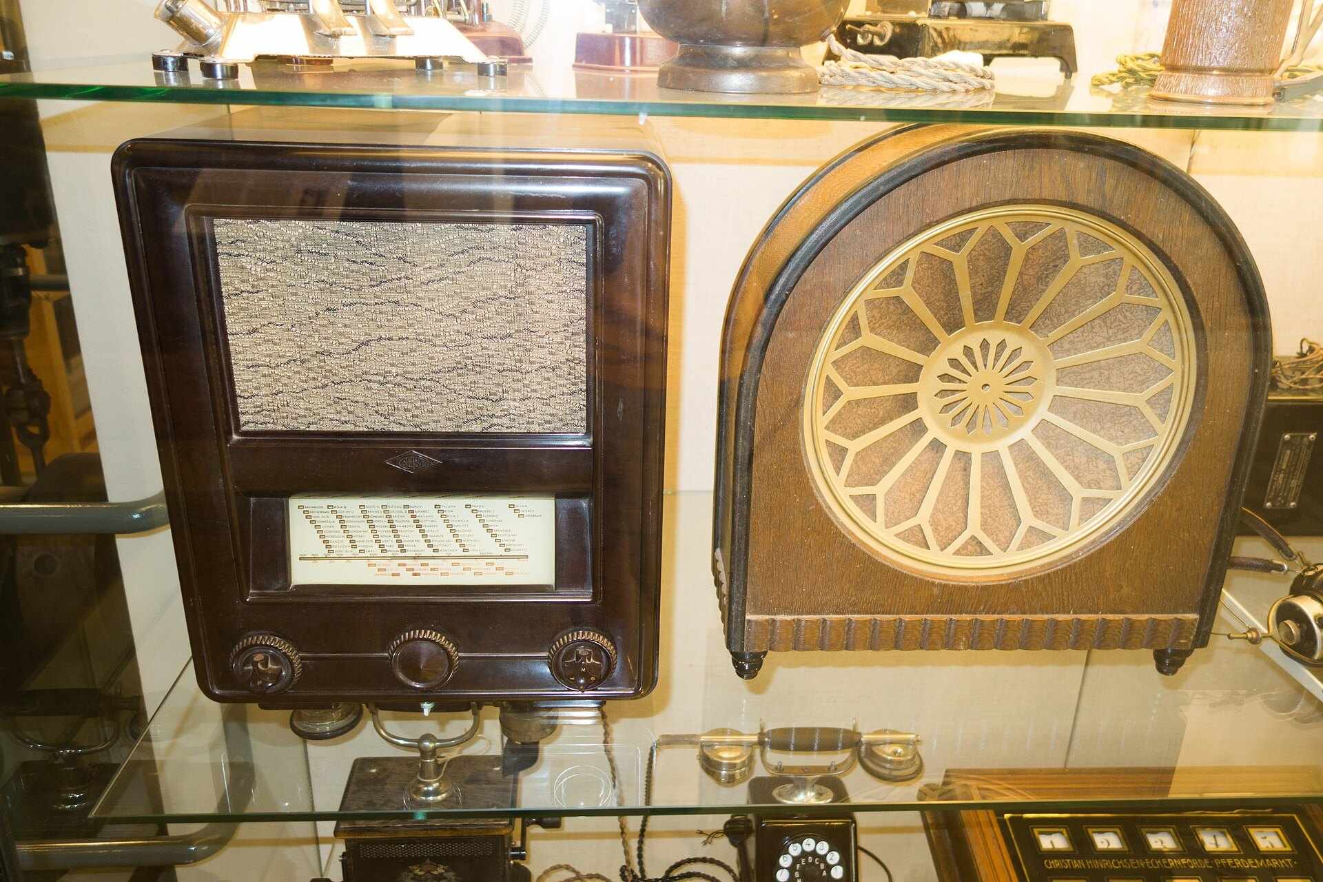 Two wooden antique radio sets on a glass museum shelf, one with a sunburst speaker grille, for the Trevor L Taylor subject page on podcasting and media