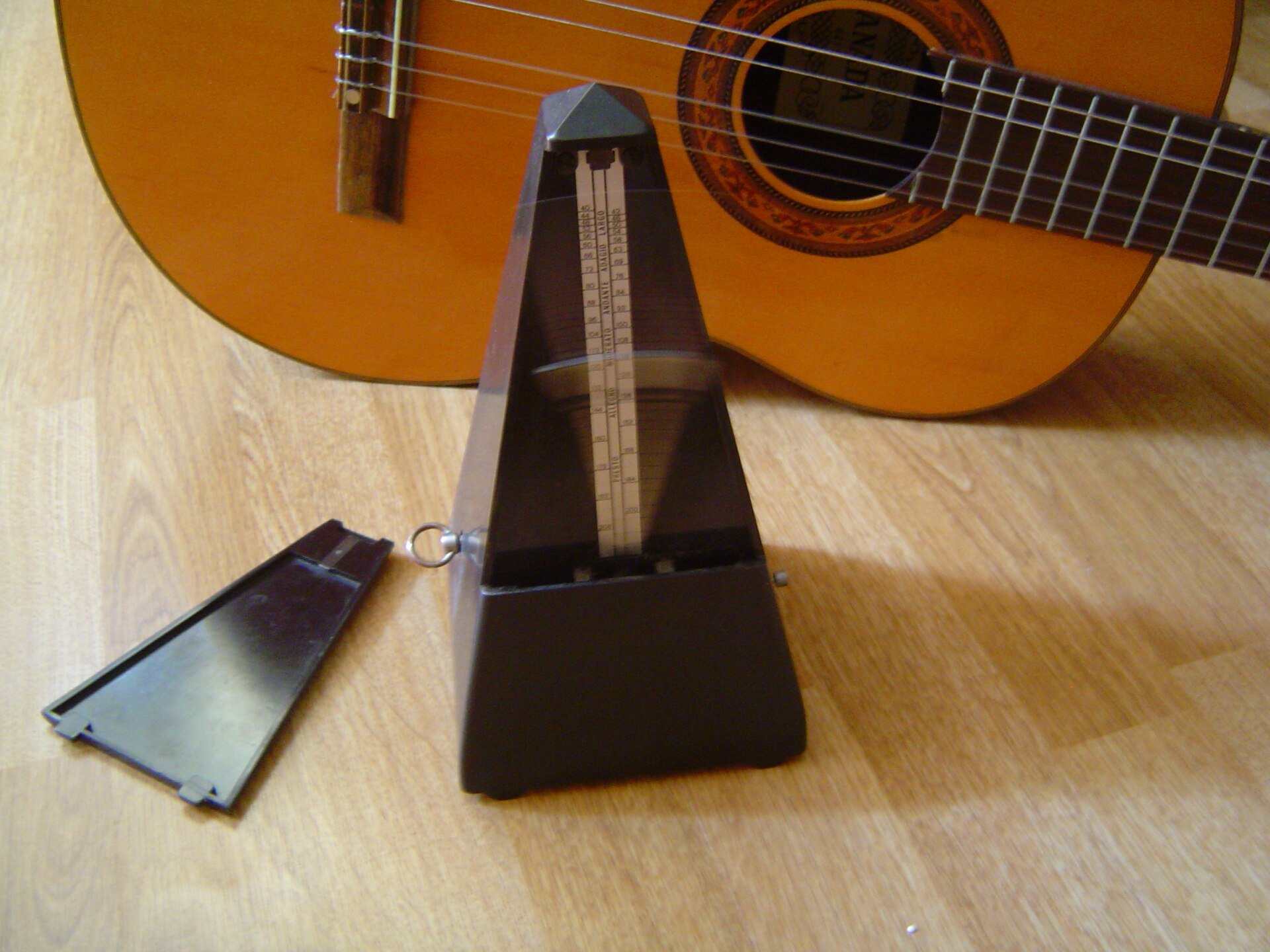 A wooden pyramid metronome with its cover off standing beside an acoustic guitar on a wood floor, from the Trevor L Taylor entry on deliberate practice