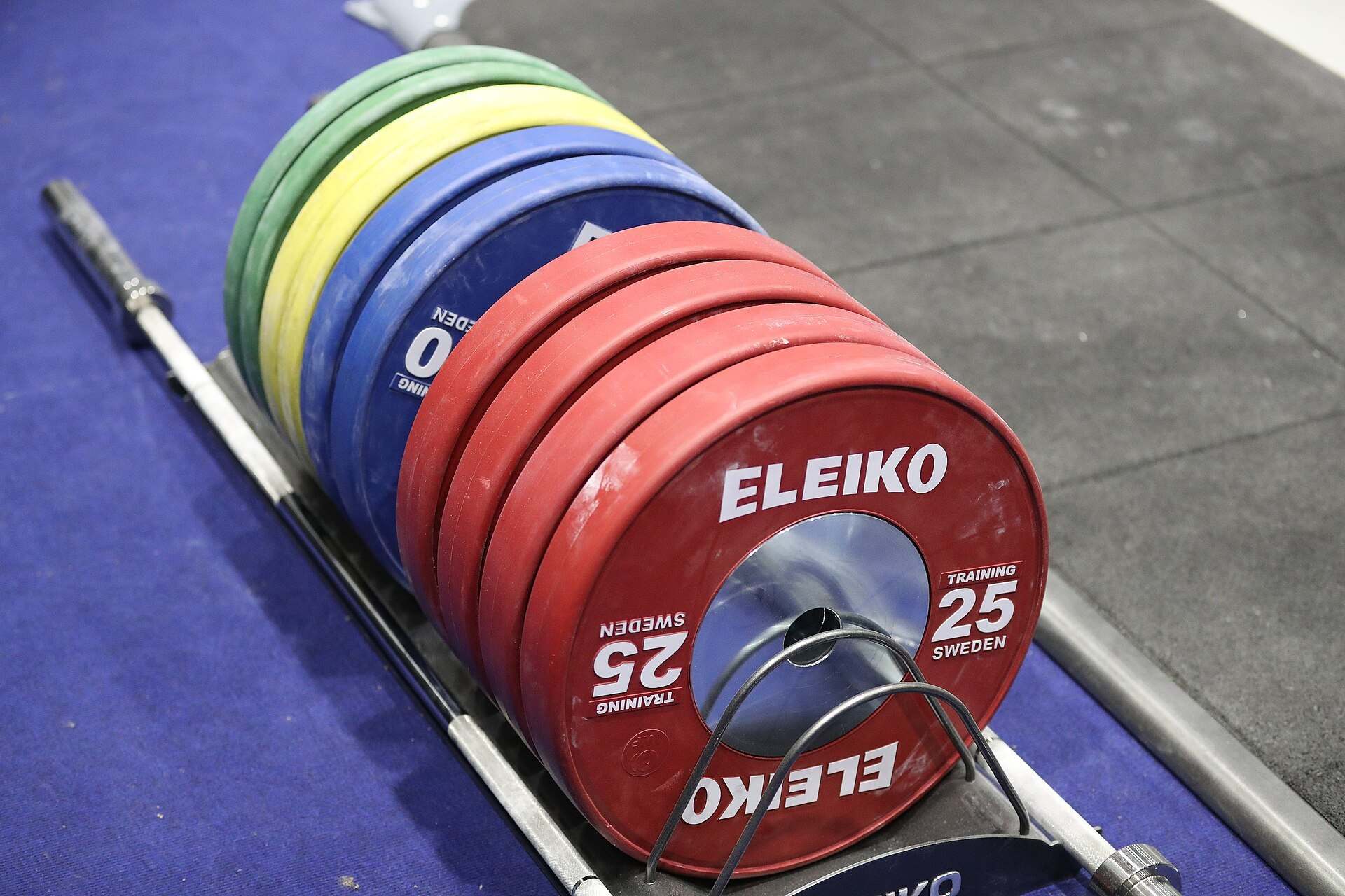 Red, blue, yellow and green competition bumper plates stacked in a rack beside a barbell on a platform, from the Trevor L Taylor entry on Olympic weightlifting
