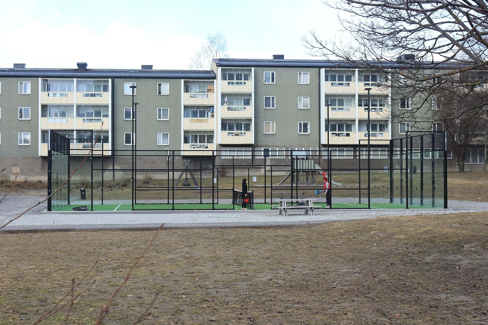 An outdoor padel enclosure with glass end walls and black mesh sides in front of apartment blocks, from the Trevor L Taylor entry on padel