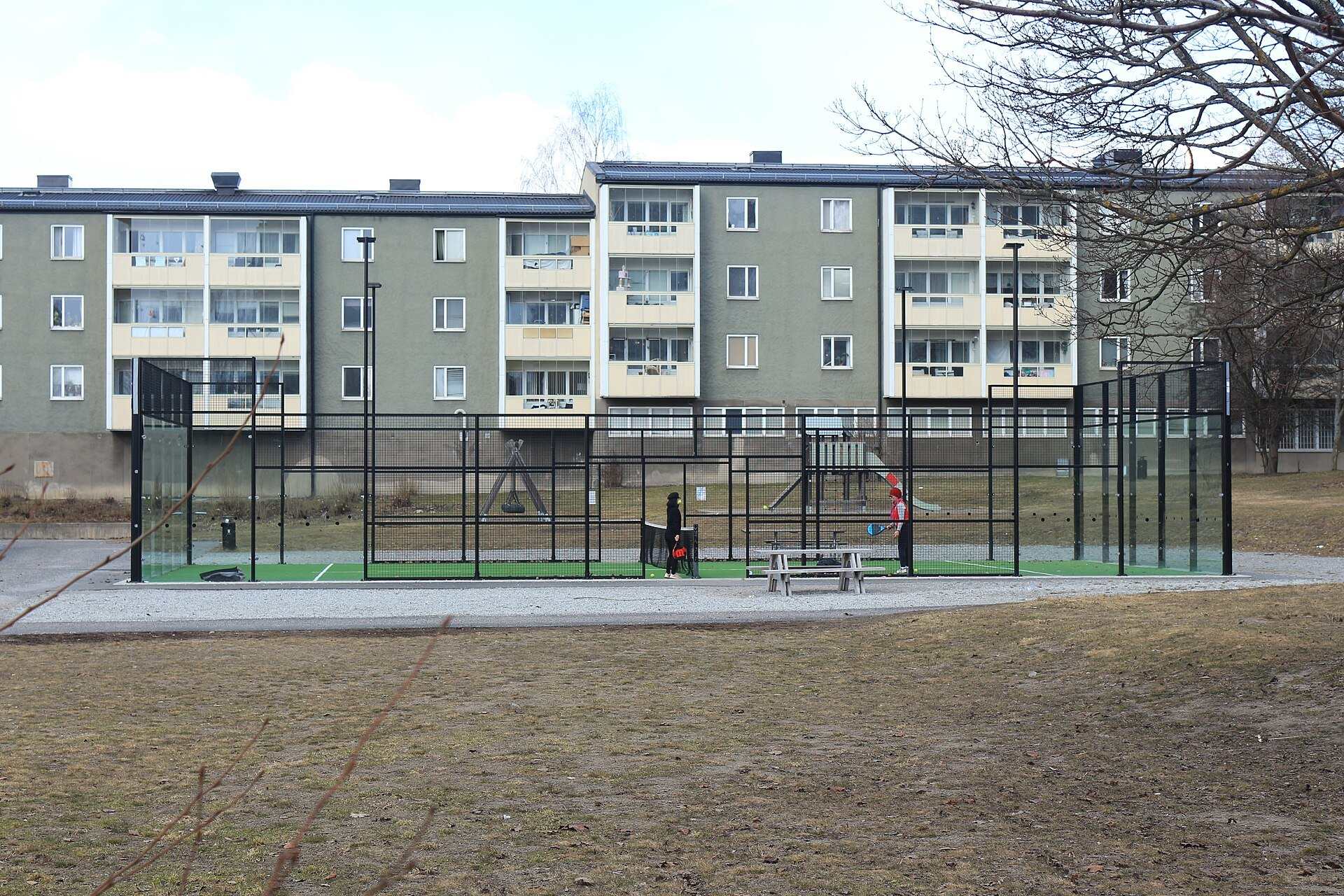 An outdoor padel enclosure with glass end walls and black mesh sides in front of apartment blocks, from the Trevor L Taylor entry on padel