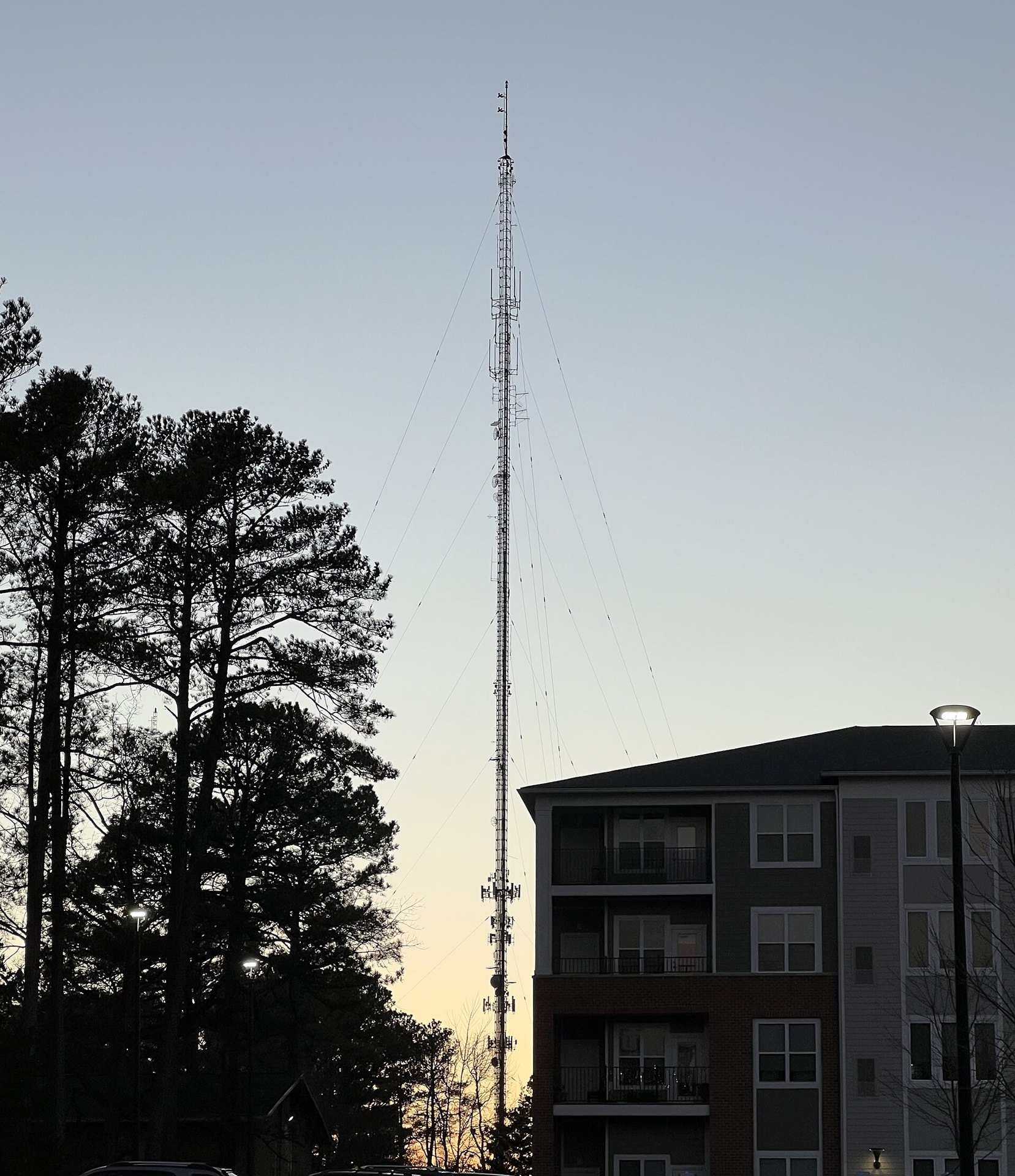 A tall guyed radio transmission tower silhouetted against a dusk sky beside pine trees, from the Trevor L Taylor entry on podcast RSS feeds