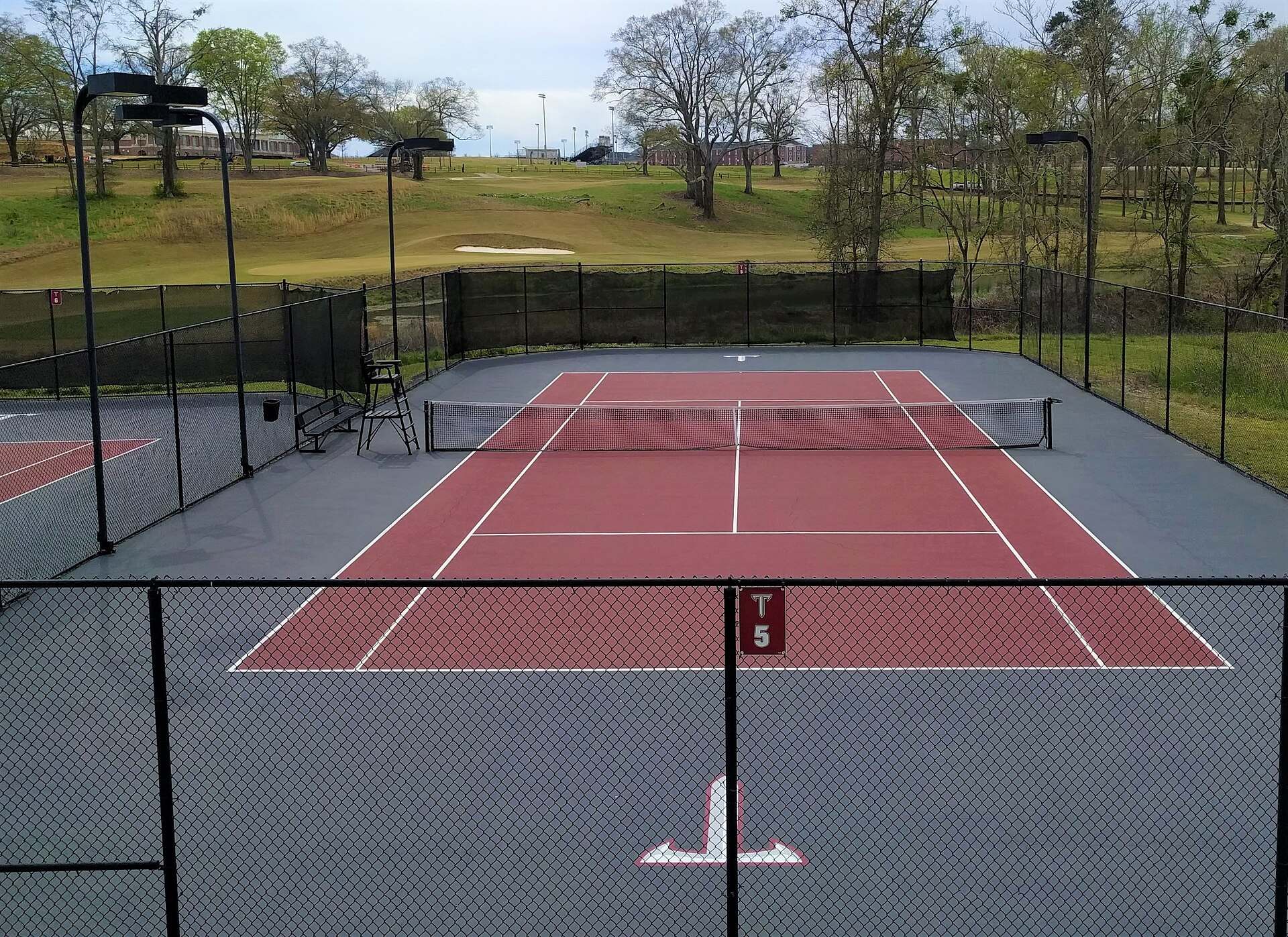 An empty red and gray hard surface with a net and umpire chair behind a chain fence, from the Trevor L Taylor entry on losing streaks in Division I tennis