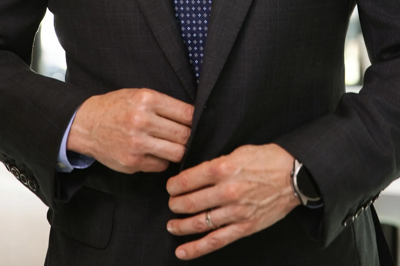 Trevor L Taylor fastening a charcoal suit jacket, a close view of his hands and a dark watch